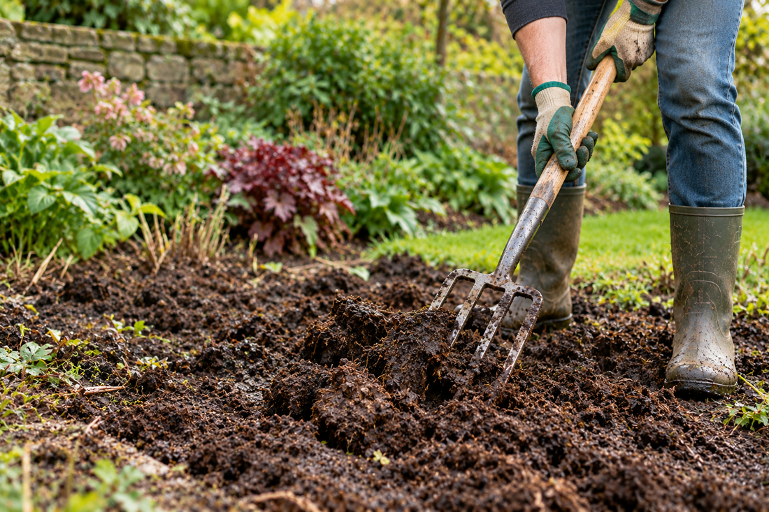 A gardener wearing rubber boots and gloves uses a garden fork to turn heavy, wet soil in a garden bed.